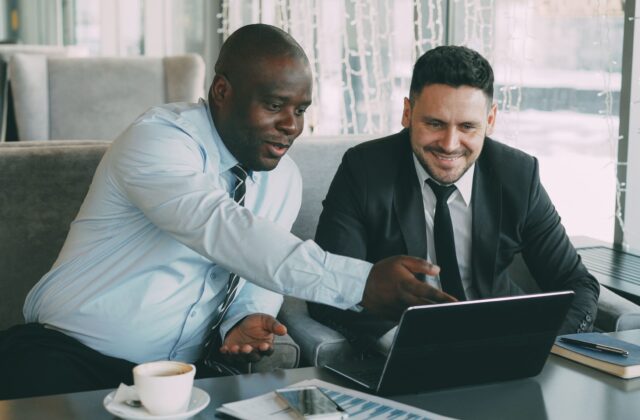 Two businessmen discussing work while using a laptop in a cafe setting.